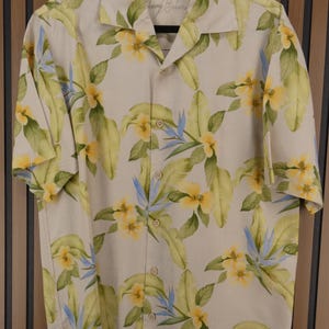 May include: Cream-colored button-down shirt with a tropical floral pattern. The shirt features yellow flowers, green leaves, and blue accents. The shirt has short sleeves and a classic collar. The brand name "Tommy Bahama" is visible on the collar.