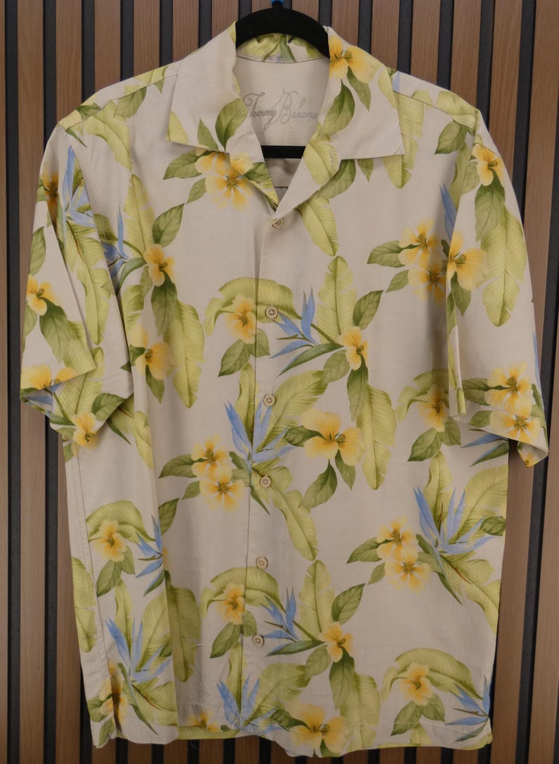 May include: Cream-colored button-down shirt with a tropical floral pattern. The shirt features yellow flowers, green leaves, and blue accents. The shirt has short sleeves and a classic collar. The brand name "Tommy Bahama" is visible on the collar.