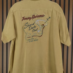 May include: Mustard yellow short-sleeved shirt with the Tommy Bahama logo and "Shark Date Club" embroidered on the back. The design includes a martini glass, shark, and anchor. The shirt has a button-down collar.