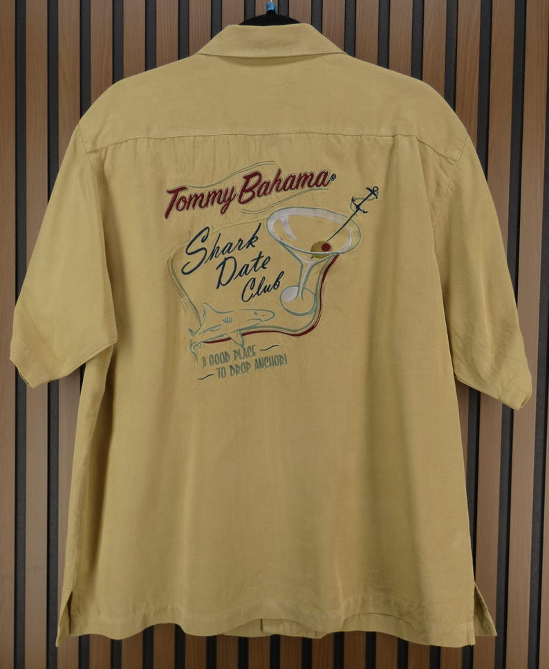 May include: Mustard yellow short-sleeved shirt with the Tommy Bahama logo and "Shark Date Club" embroidered on the back. The design includes a martini glass, shark, and anchor. The shirt has a button-down collar.