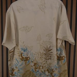 K&ouml;nnte beinhalten: Cremefarbenes Button-Down-Hemd mit tropischem Blatt- und Blumenmuster in Braun-, Blau- und Gr&uuml;nt&ouml;nen. Das kurz&auml;rmelige Hemd h&auml;ngt an einem schwarzen Kleiderb&uuml;gel vor einem Holzgrund.