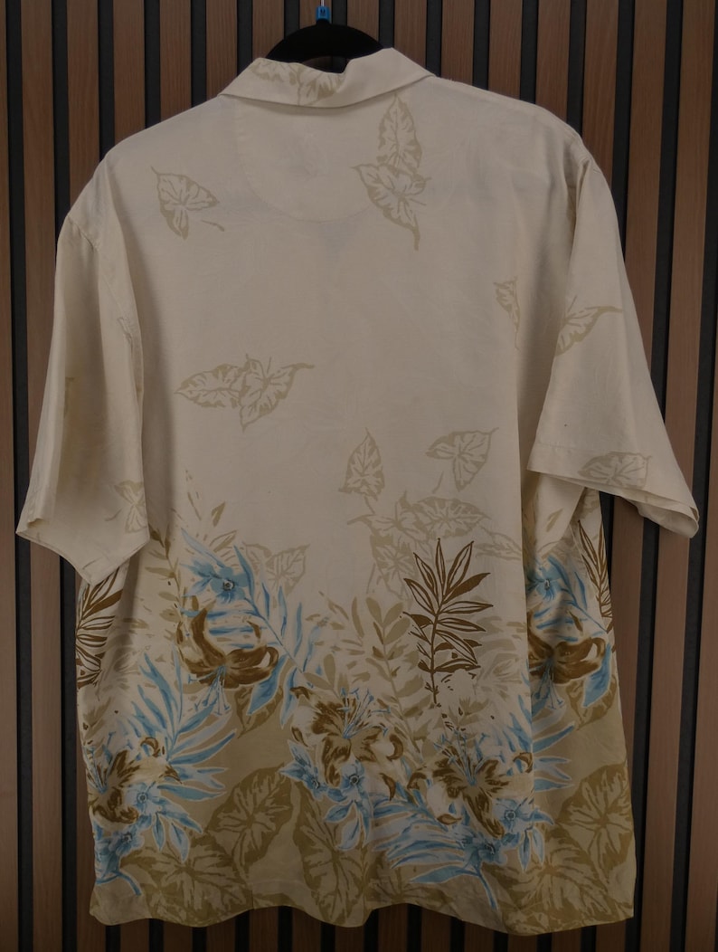 K&ouml;nnte beinhalten: Cremefarbenes Button-Down-Hemd mit tropischem Blatt- und Blumenmuster in Braun-, Blau- und Gr&uuml;nt&ouml;nen. Das kurz&auml;rmelige Hemd h&auml;ngt an einem schwarzen Kleiderb&uuml;gel vor einem Holzgrund.