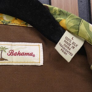 May include: Close-up of a Tommy Bahama silk shirt label. The label reads "Tommy Bahama" with a palm tree logo. The shirt is brown with a colorful floral pattern on the collar. The tag reads "100% Silk Made in China Copyrighted Print."
