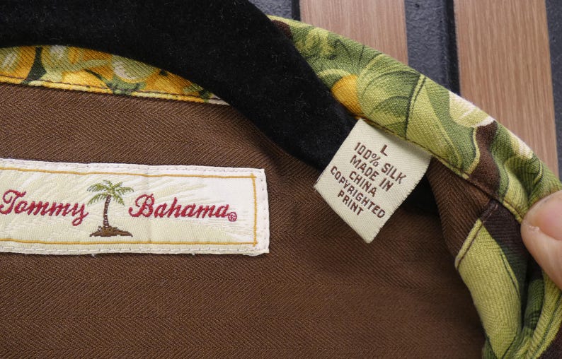 May include: Close-up of a Tommy Bahama silk shirt label. The label reads "Tommy Bahama" with a palm tree logo. The shirt is brown with a colorful floral pattern on the collar. The tag reads "100% Silk Made in China Copyrighted Print."