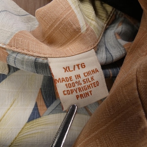 Puede incluir: Primer plano de una camisa de seda con un estampado floral colorido. La etiqueta de la camisa dice "XL/TG MADE IN CHINA 100% SILK COPYRIGHTED PRINT". La tela presenta tonos beige, azul y verde.