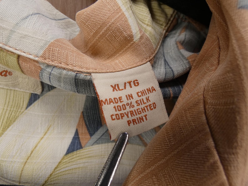 Puede incluir: Primer plano de una camisa de seda con un estampado floral colorido. La etiqueta de la camisa dice "XL/TG MADE IN CHINA 100% SILK COPYRIGHTED PRINT". La tela presenta tonos beige, azul y verde.
