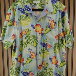 May include: Light blue button-down shirt with a tropical floral pattern. The shirt features a vibrant design of yellow, red, and orange flowers, along with green and blue leaves. The shirt has short sleeves and a button-up front. The brand name "Tommy Bahama" is visible on the collar.