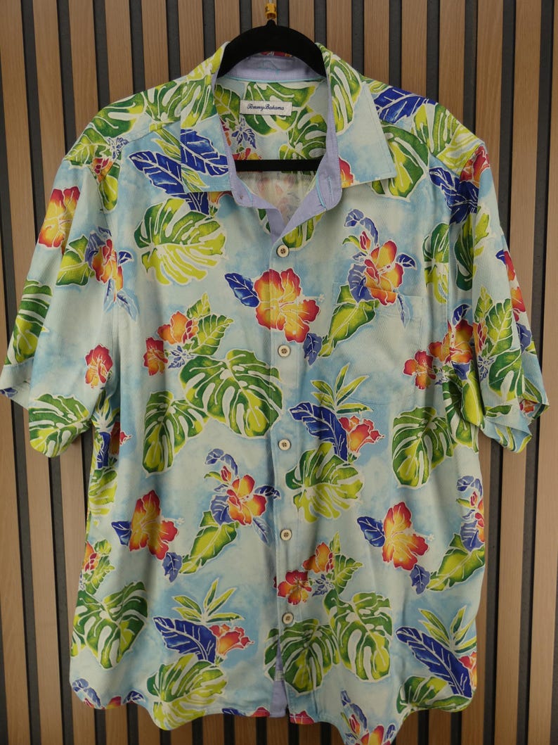 May include: Light blue button-down shirt with a tropical floral pattern. The shirt features a vibrant design of yellow, red, and orange flowers, along with green and blue leaves. The shirt has short sleeves and a button-up front. The brand name "Tommy Bahama" is visible on the collar.