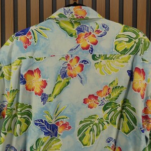May include: Light blue button-down shirt with a vibrant floral pattern. The design features red, yellow, and orange hibiscus flowers, green leaves, and blue accents. The shirt is made of a lightweight fabric, perfect for warm weather.