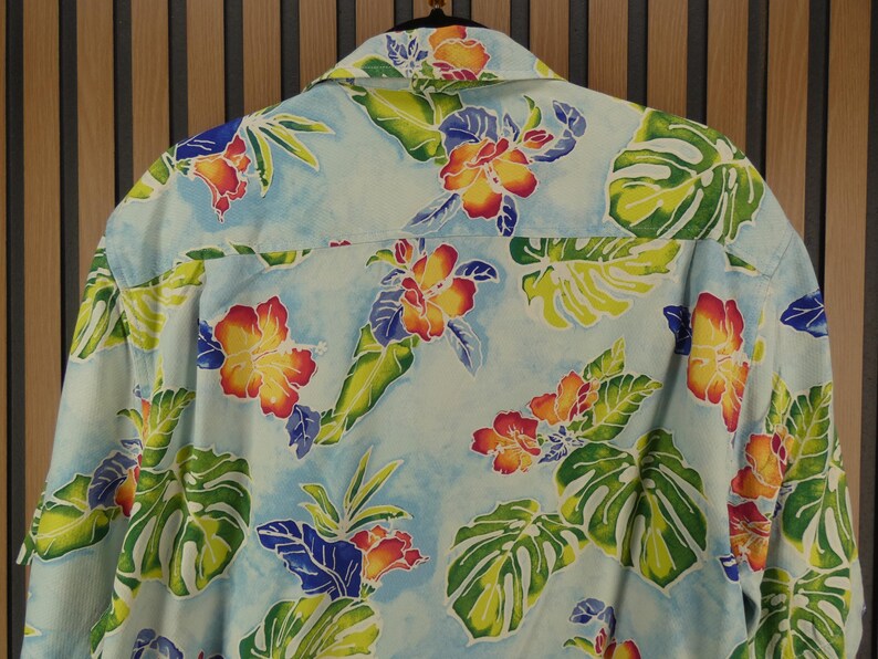 May include: Light blue button-down shirt with a vibrant floral pattern. The design features red, yellow, and orange hibiscus flowers, green leaves, and blue accents. The shirt is made of a lightweight fabric, perfect for warm weather.