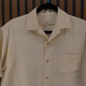 May include: Cream-colored button-down shirt with short sleeves, a chest pocket, and a collar. The shirt has the brand name "Tommy Bahama" embroidered on the collar. The shirt is hanging on a black hanger.