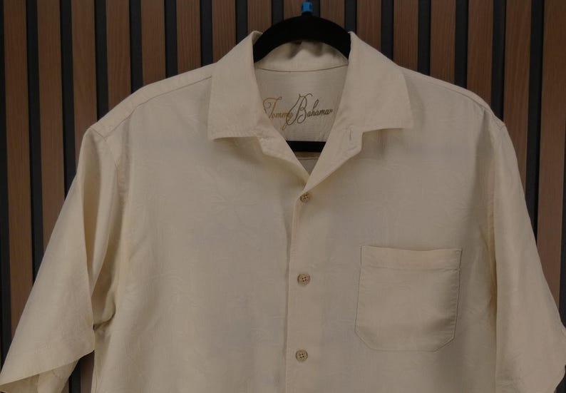 May include: Cream-colored button-down shirt with short sleeves, a chest pocket, and a collar. The shirt has the brand name "Tommy Bahama" embroidered on the collar. The shirt is hanging on a black hanger.