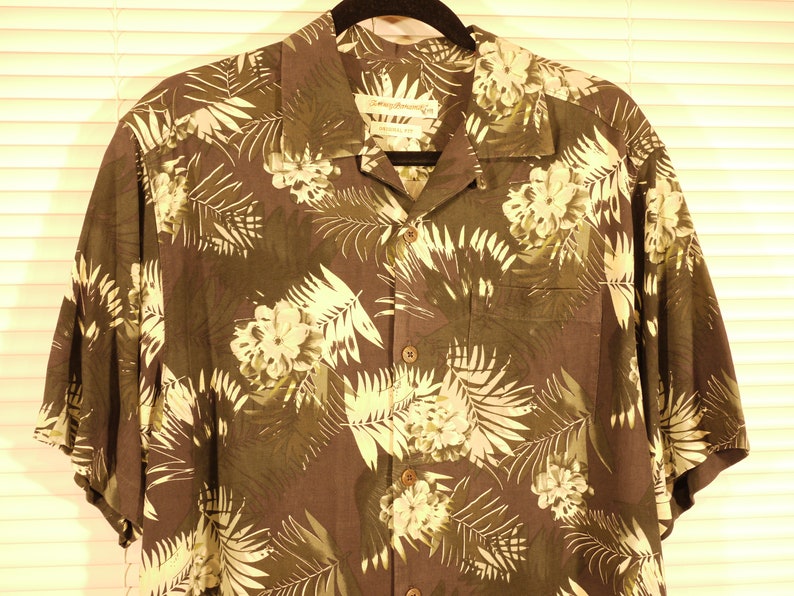 May include: A dark brown short-sleeved button-down shirt with a tropical print featuring white and green leaves and flowers. The shirt has a collar and a pocket on the left side.
