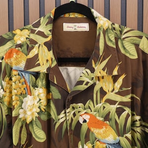 May include: Brown button-up shirt with a tropical parrot and floral print. The shirt features a collar and short sleeves. The design includes parrots, yellow flowers, and green leaves. The shirt is hanging on a black hanger.