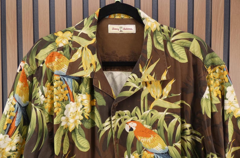May include: Brown button-up shirt with a tropical parrot and floral print. The shirt features a collar and short sleeves. The design includes parrots, yellow flowers, and green leaves. The shirt is hanging on a black hanger.