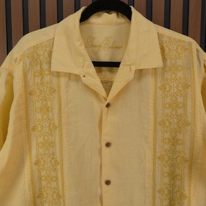 May include: A pale yellow short-sleeved shirt with a button-down front and a collar. The shirt has embroidered floral patterns in a darker yellow shade down the front. The brand name "Tommy Bahama" is embroidered on the collar.