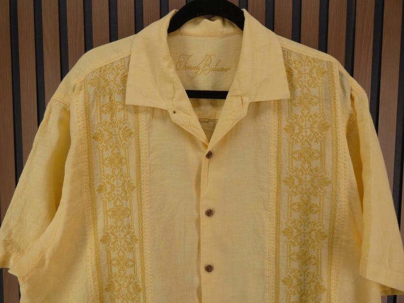 May include: A pale yellow short-sleeved shirt with a button-down front and a collar. The shirt has embroidered floral patterns in a darker yellow shade down the front. The brand name "Tommy Bahama" is embroidered on the collar.
