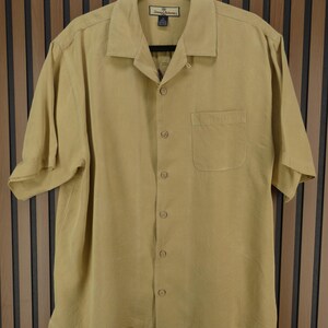 May include: A short-sleeved, button-down shirt in a light tan color. The shirt has a collared neckline, a single chest pocket, and a relaxed fit. The shirt is hanging on a black hanger.