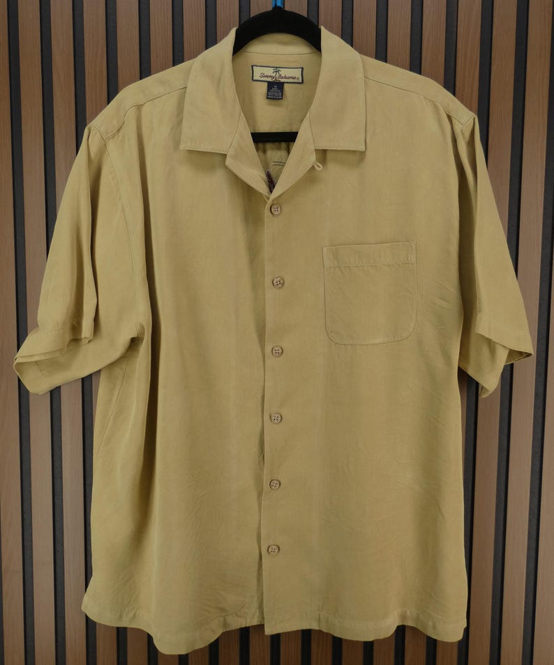 May include: A short-sleeved, button-down shirt in a light tan color. The shirt has a collared neckline, a single chest pocket, and a relaxed fit. The shirt is hanging on a black hanger.