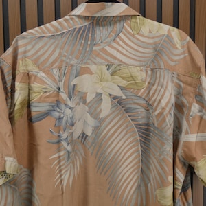 Puede incluir: Una camisa de manga corta con un estampado floral tropical. La camisa presenta una base de color melocot&oacute;n con grandes hojas y flores estilizadas en tonos de blanco, gris y amarillo. Cuelga sobre un fondo de madera.