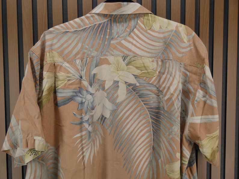 Puede incluir: Una camisa de manga corta con un estampado floral tropical. La camisa presenta una base de color melocot&oacute;n con grandes hojas y flores estilizadas en tonos de blanco, gris y amarillo. Cuelga sobre un fondo de madera.