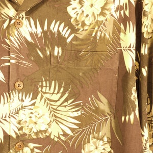 May include: A brown button-up shirt with a tropical print featuring white palm leaves and flowers. The shirt has a pocket on the left side and two buttons on the front.