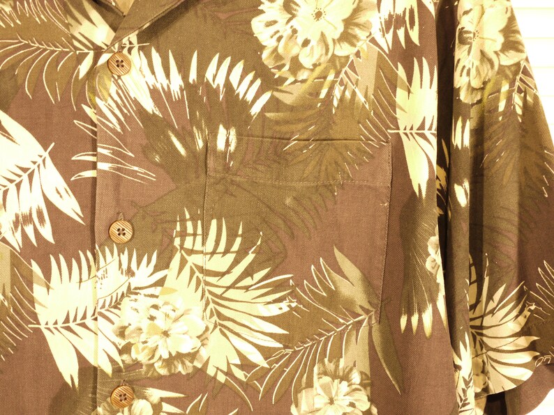 May include: A brown button-up shirt with a tropical print featuring white palm leaves and flowers. The shirt has a pocket on the left side and two buttons on the front.