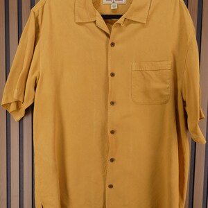 May include: Mustard yellow short-sleeve button-down shirt with a single chest pocket. The shirt has a collared neckline and is hanging on a black hanger. The brand name is visible on the label.
