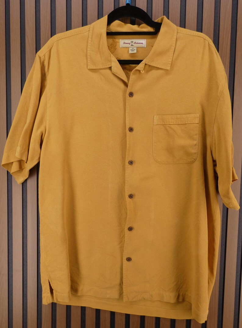 May include: Mustard yellow short-sleeve button-down shirt with a single chest pocket. The shirt has a collared neckline and is hanging on a black hanger. The brand name is visible on the label.