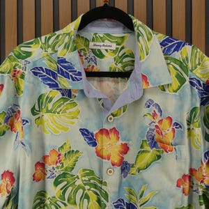 May include: Light blue button-down shirt with a tropical floral pattern. The shirt features large green leaves, red, yellow, and orange flowers, and blue accents. The shirt is hanging on a black hanger.