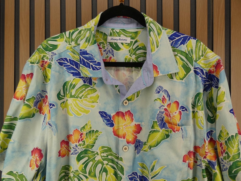 May include: Light blue button-down shirt with a tropical floral pattern. The shirt features large green leaves, red, yellow, and orange flowers, and blue accents. The shirt is hanging on a black hanger.