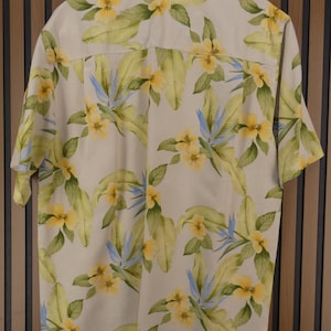 May include: A short-sleeved button-down shirt with a cream-colored background. The shirt features a vibrant floral pattern with yellow flowers, green leaves, and blue accents. The shirt is made of a lightweight fabric.