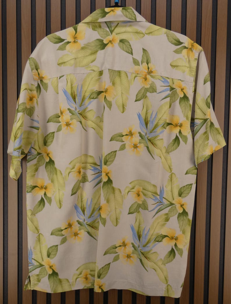 May include: A short-sleeved button-down shirt with a cream-colored background. The shirt features a vibrant floral pattern with yellow flowers, green leaves, and blue accents. The shirt is made of a lightweight fabric.