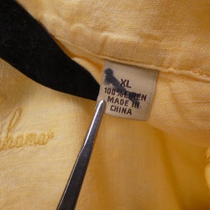 May include: Close-up of a yellow linen shirt with the embroidered word "Bahama" in a cursive font. The shirt tag reads "XL 100% LINEN MADE IN CHINA". The shirt is hanging on a black hanger.