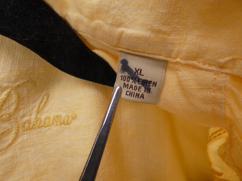 May include: Close-up of a yellow linen shirt with the embroidered word "Bahama" in a cursive font. The shirt tag reads "XL 100% LINEN MADE IN CHINA". The shirt is hanging on a black hanger.