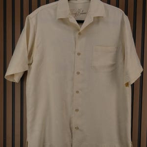 May include: Cream-colored short-sleeve button-down shirt with a chest pocket. The shirt has a classic collar and is hanging on a black hanger. The brand name "Tommy Bahama" is visible on the collar.