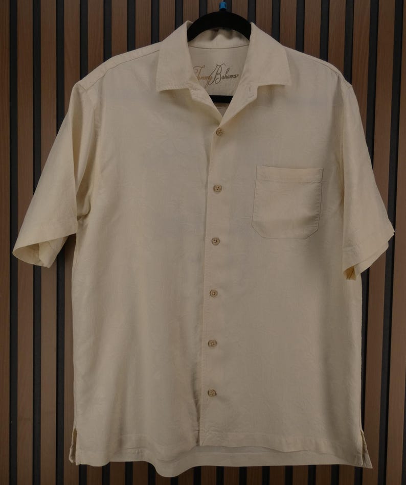 May include: Cream-colored short-sleeve button-down shirt with a chest pocket. The shirt has a classic collar and is hanging on a black hanger. The brand name "Tommy Bahama" is visible on the collar.