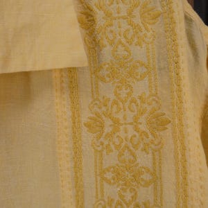 May include: A close-up of a pale yellow shirt with detailed yellow embroidery. The embroidery showcases a floral and geometric design down the centre and edges. The fabric appears to be linen or a similar lightweight material, ideal for warmer weather.