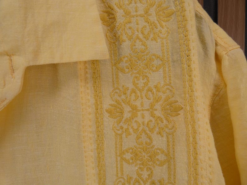 May include: A close-up of a pale yellow shirt with detailed yellow embroidery. The embroidery showcases a floral and geometric design down the centre and edges. The fabric appears to be linen or a similar lightweight material, ideal for warmer weather.
