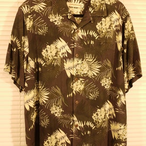 May include: A dark green short-sleeved button-down shirt with a tropical leaf and flower print. The shirt has a collar and a pocket on the left side. The label reads "Island Traders".