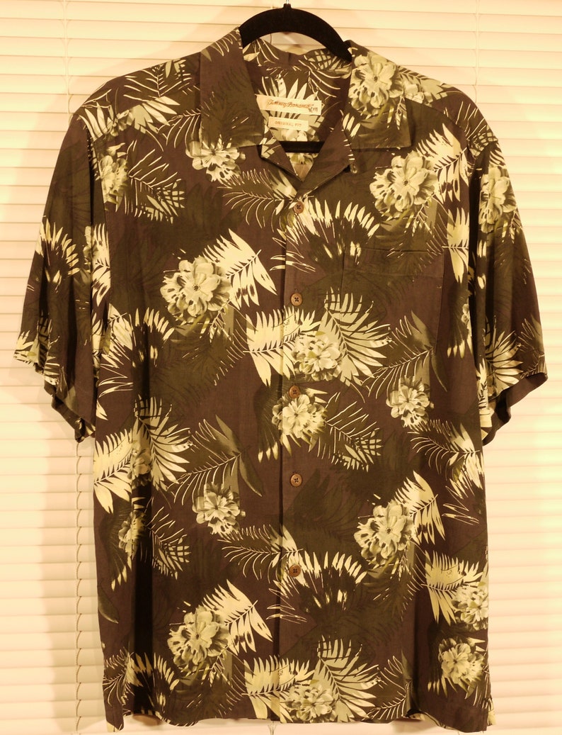 May include: A dark green short-sleeved button-down shirt with a tropical leaf and flower print. The shirt has a collar and a pocket on the left side. The label reads "Island Traders".