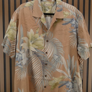 Puede incluir: Una camisa de manga corta con botones de color melocot&oacute;n, adornada con un estampado floral tropical en tonos blancos, azules y amarillos. La camisa est&aacute; colgada de una percha negra.