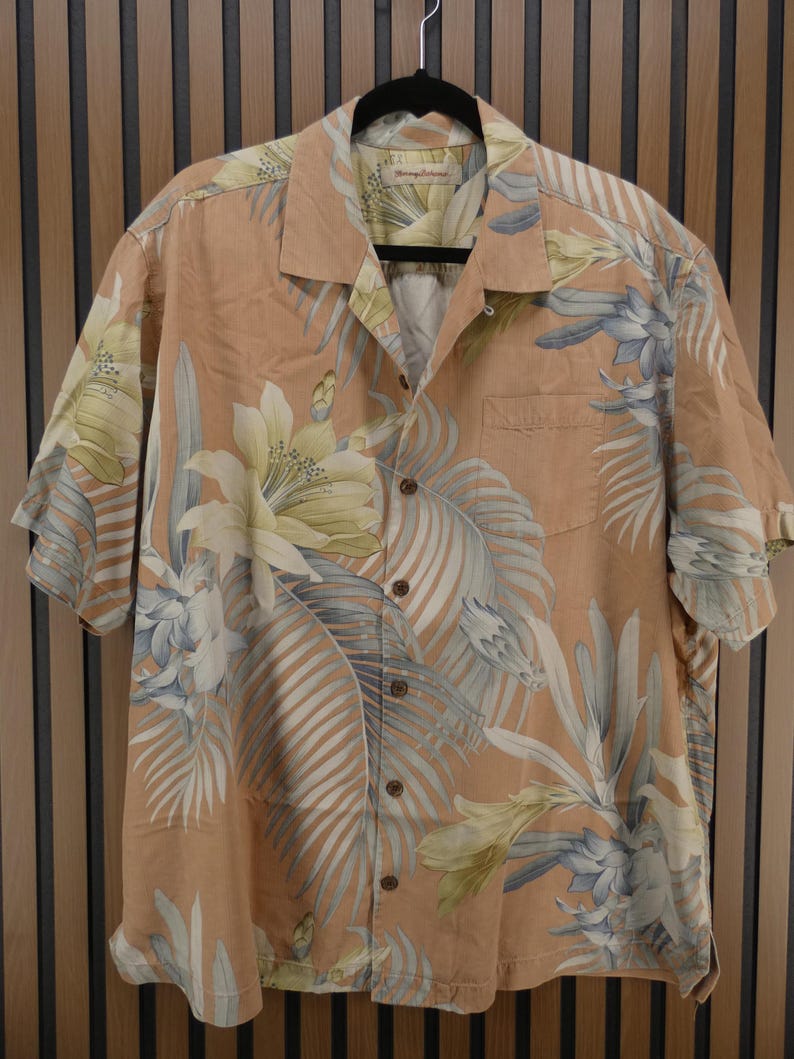 Puede incluir: Una camisa de manga corta con botones de color melocot&oacute;n, adornada con un estampado floral tropical en tonos blancos, azules y amarillos. La camisa est&aacute; colgada de una percha negra.