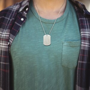 Puede incluir: Un collar de plata con un colgante de placa de identificaci&oacute;n que tiene la Oraci&oacute;n de la Serenidad grabada. El collar lo lleva una persona que lleva una camiseta verde y una camisa de cuadros.