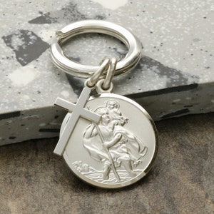 May include: Silver keyring featuring a Saint Christopher medal with a cross charm. The medal depicts Saint Christopher carrying a child on his shoulder, walking through water.