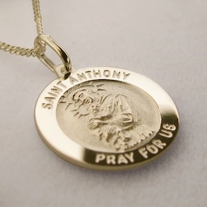 May include: Gold pendant necklace with a round charm featuring a depiction of Saint Anthony holding a baby. The charm is inscribed with "Saint Anthony Pray For Us".