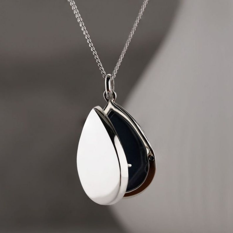May include: A silver teardrop-shaped locket necklace with a hinged opening. The locket is open, revealing a black interior.