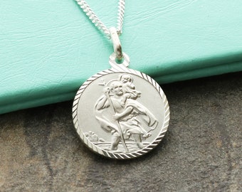 May include: Silver chain necklace with a round pendant featuring a detailed image of Saint Christopher carrying a child. The pendant has a textured edge.
