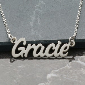 May include: A silver necklace featuring the name "Gracie" in a cursive script. The pendant hangs from a fine chain. The necklace is presented against a dark grey backdrop.