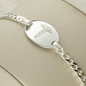 Puede incluir: Un brazalete de identificaci&oacute;n m&eacute;dica de plata con un dise&ntilde;o de cadena de eslabones y una etiqueta ovalada redonda grabada con el s&iacute;mbolo m&eacute;dico, el bast&oacute;n de Asclepio.
